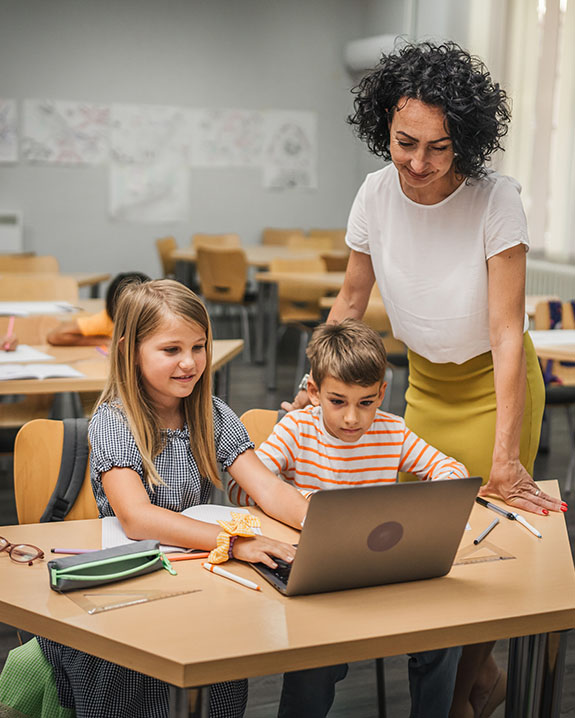 Two students sitting at desk looking at a laptop, the teacher is standing behind them