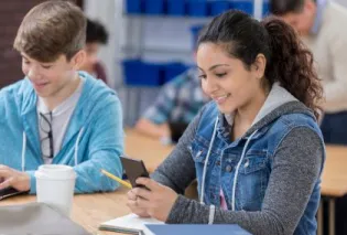two high school students holding cell phone and typing on computer