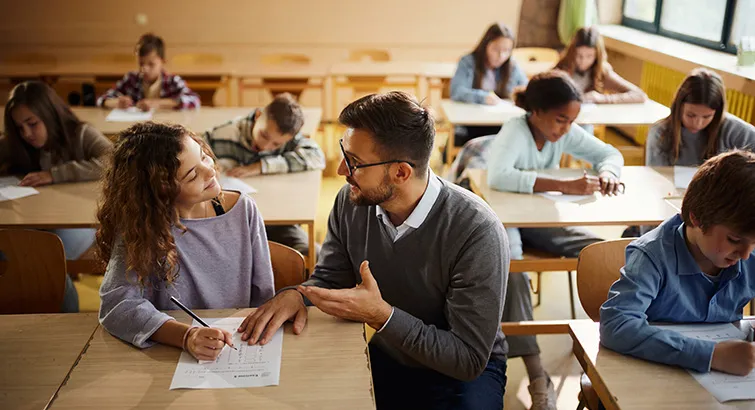 Male teacher kneeling down to work with young student at her desk