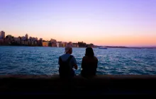 Two people sitting by the water looking out over a sunset