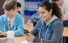 two high school students holding cell phone and typing on computer