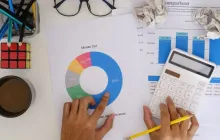 Desk from above with papers containing graphs and numbers, hands pointing to graphs, calculator
